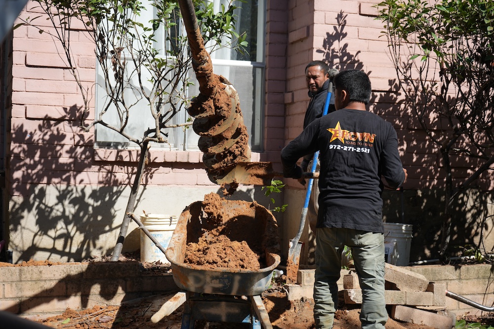 Brown Foundation Repair crew drilling a hole for a concrete pier using an auger, collecting the soil in a wheelbarrow for drilled pier foundation repair.